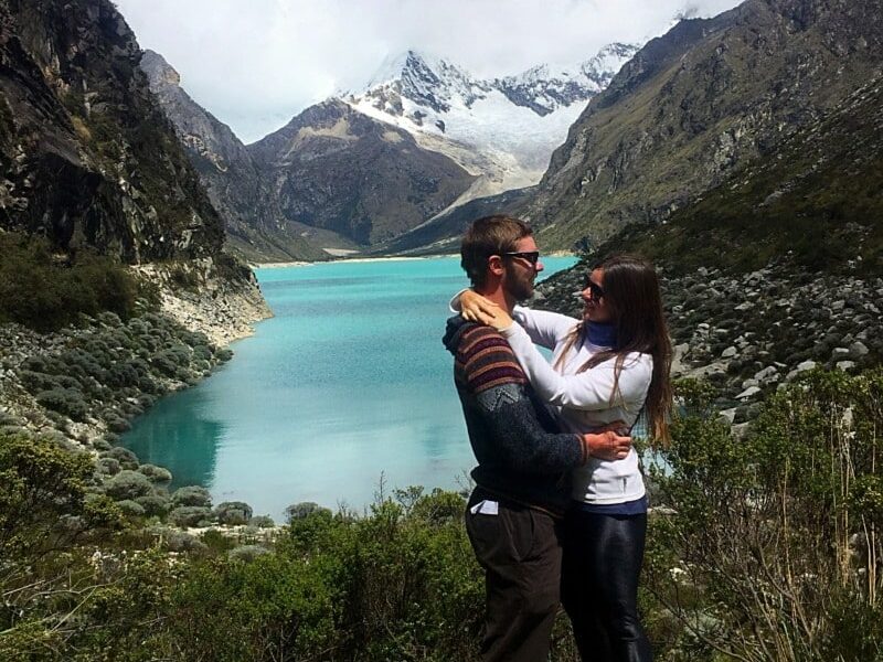 Casal abraçados e sorrindo, com a vista de um lago cristalino azul rodeado de montanhas cobertas de natureza e neve atrás. Representa o que fazer no Peru