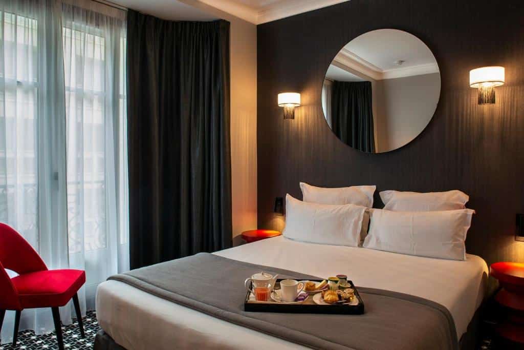 Quarto do Lenox Montparnasse, um dos hotéis 3 estrelas em Paris. Uma cama de casal está encostada na parede ao lado direito com mesinhas de cabeceira e abajures dos dois lados, assim como um espelho na parede acima e uma bandeja com café da manhã aos pés. Na lateral esquerda do quarto há uma janela com cortinas e uma poltrona.