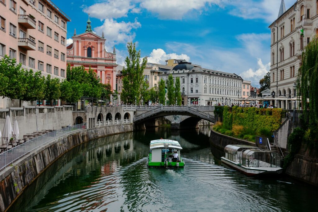 Cena urbana no centro de Liubliana, Eslovênia. O Rio Ljubljanica flui calmamente através da cidade, ladeado por edifícios históricos coloridos e uma igreja com uma fachada rosa marcante. Uma ponte de pedra ornamentada cruza o rio, enquanto pessoas passeiam e desfrutam da atmosfera da cidade. Um barco turístico navega suavemente pelo rio, adicionando movimento à paisagem tranquila e bem cuidada que reflete a arquitetura e a vida ao redor.
