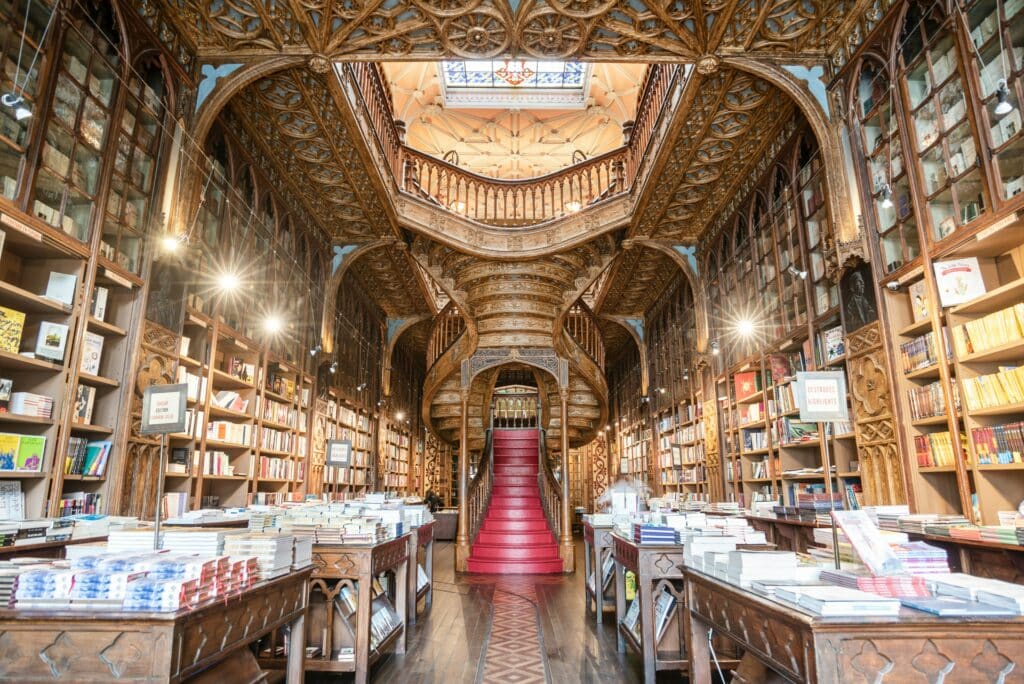 Imagem da Livraria Lello por dentro a frente bancas de livros em cada lado da imagem e ao fundo no centro a escadaria que leva acesso ao andar de cima.