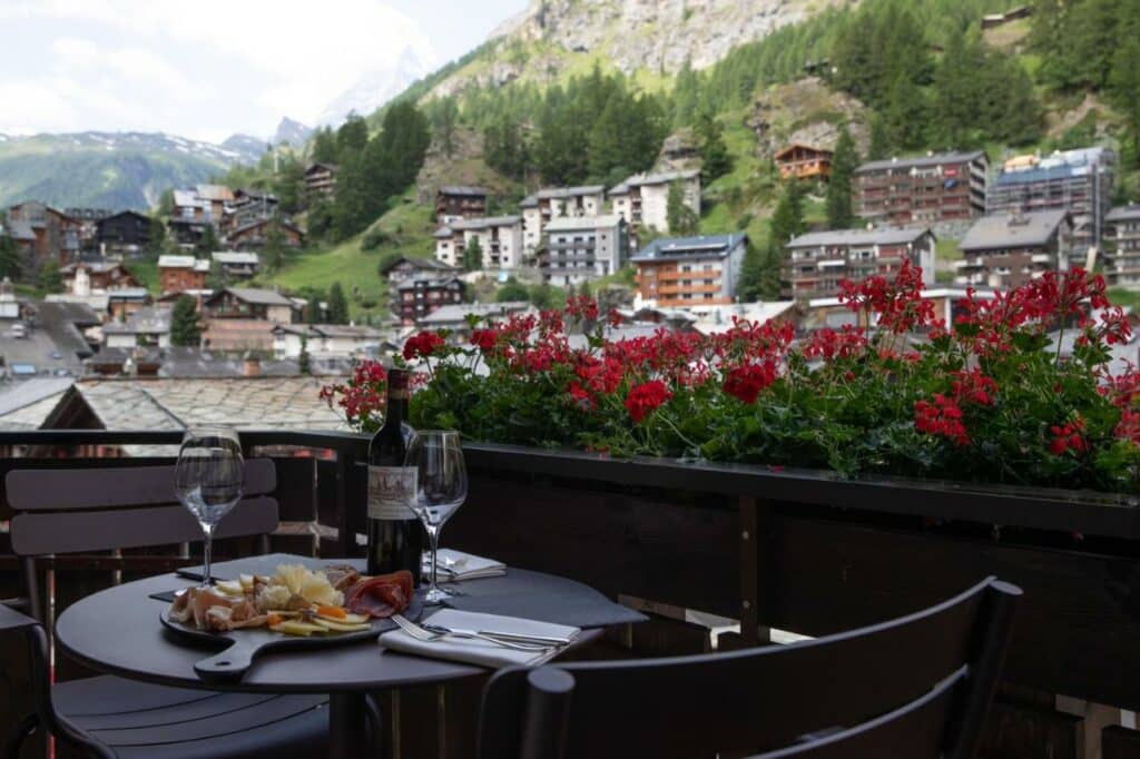 Varanda do Schweizerhof Zermatt – a Small Luxury Hotel durante o dia com uma mesa redonda do no lado esquerdo com três cadeiras e em cima da mesa duas taças, com vinho e tabuas de frios com vista para a cidade.
