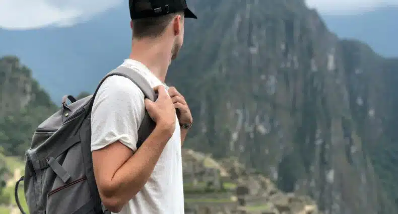 Homem de blusa branca e boné preto, olhando para a direita, com uma mochila nas costas e olhando para a montanha e cidade perdida de Machu Picchu. Representa o que levar na mala para o Peru