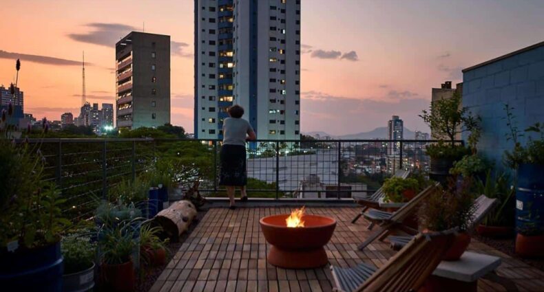 A imagem mostra o terraço da Vila Pitanga Hosting, um dos airbnb em São Paulo. Em primeiro plano, uma fogueira de chão acesa cria um ambiente acolhedor no centro do espaço. Duas cadeiras de madeira tipo espreguiçadeira estão posicionadas ao redor da fogueira, prontas para relaxamento. O deck de madeira é ladeado por uma variedade de plantas em vasos, adicionando toques de verde à cena. Ao fundo, uma mulher de costas está apreciando a vista da cidade, com prédios altos e luzes começando a brilhar sob um céu em tons de roxo e laranja que anunciam o final do dia. A atmosfera é tranquila e contemplativa.