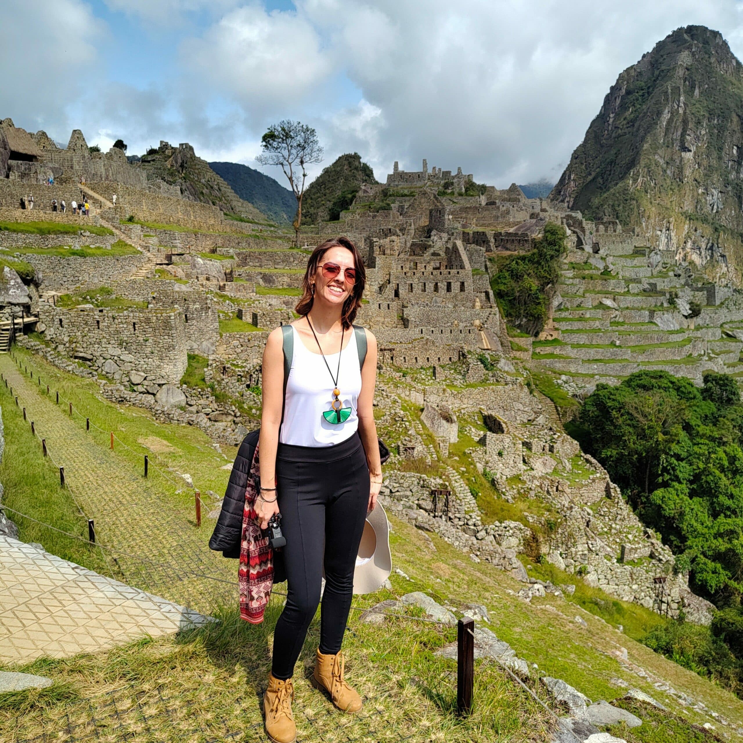 Mulher sorrindo, com mochila nas costas e a vista de Machu Picchu ao fundo. Representa o que levar para o Peru