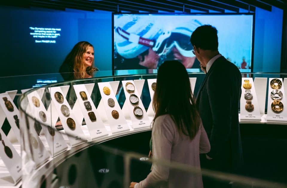 Imagem do Museu Olímpico com duas pessoas olhando a exposição de medalhas e atrás uma mulher.