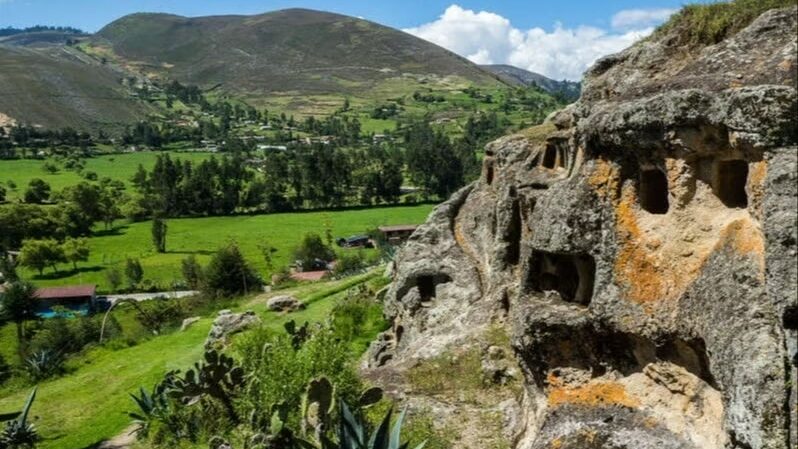Nécropole de Utuzco. Na imagem, há uma montanha de pedras à direita, e um campo com gramado verde, árvores e montanhas em volta. Foto: Civitatis