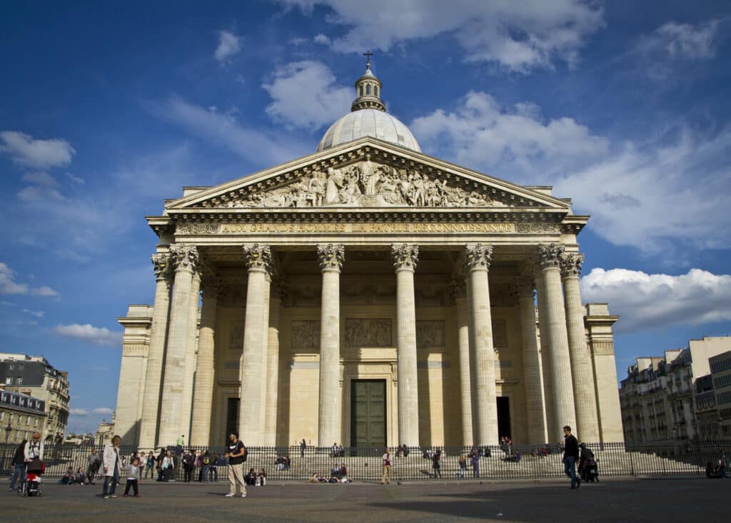 Fachada do Panteão, uma das opções de o que fazer em Paris. Várias colunas e figuras romanas fazem parte do local, e há um domo no topo. Há algumas pessoas passeando ao redor.