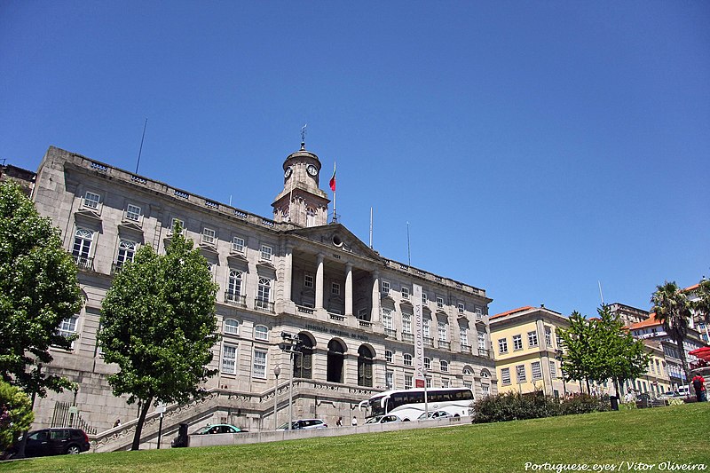 Imagem do Palácio da Bolsa durante o dia com um jardim a frente do lado esquerdo carros parados e atrás o edifício. Representa o que fazer no Porto.