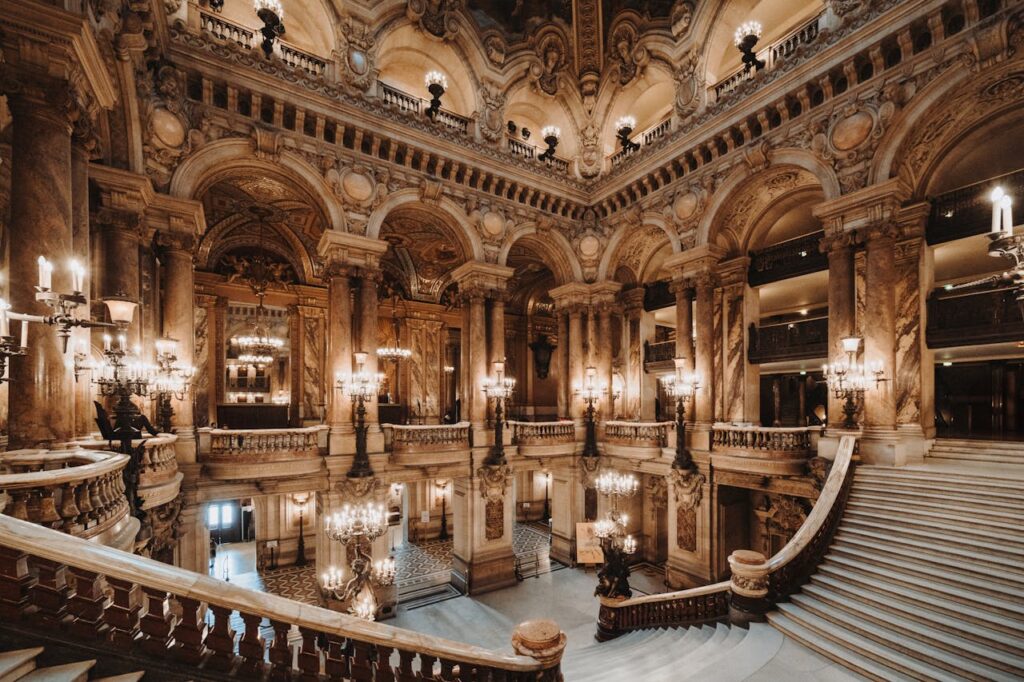Escadarias e colunas rebuscadas na Ópera Garnier, uma das opções de o que fazer em Paris. Lustres e luminárias luxuosas estçao espalhadas pelo local.
