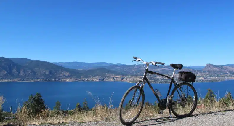 Uma bicicleta está estacionada em um caminho de cascalho com vista para um grande lago azul e colinas distantes sob um céu azul claro, no Okanagan.