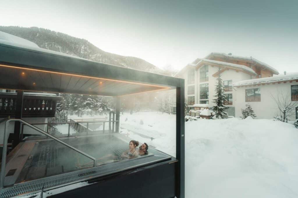 Uma banheira de hidromassagem do lado esquerdo com um casal dentro no Alpen Resort & Spa durante o dia com vista para a neve e ao fundo a estadia.