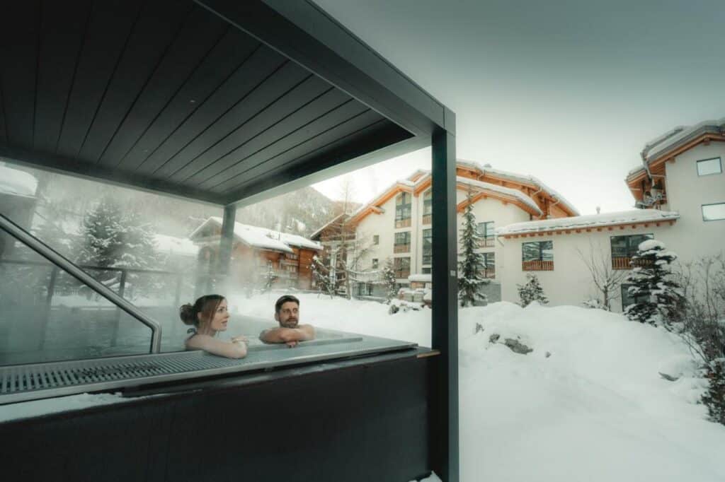 Um casal dentro da banheira de hidromassagem no Zermatt Budget Rooms durante o dia com banheira do lado esquerdo da imagem do lado direito jardim coberto com neve e a estadia.