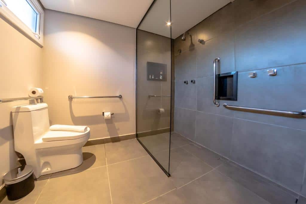 Banheiro de um dos quartos da Pousada Boutique Figueira da Serra. No lado esquerdo está a privada do banheiro onde há duas barras de apoio. Já no lado direito há o box onde está o chuveiro e outras duas barras de apoio.