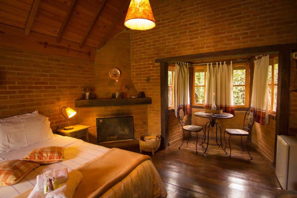 Quarto da Pousada Canto da Lua. Uma cama de casal com uma estante e um abajur do lado esquerdo, no fundo uma lareira, janelas e no meio uma mesa com duas cadeiras. Foto para ilustrar post sobre pousadas pet friendly em São Paulo.