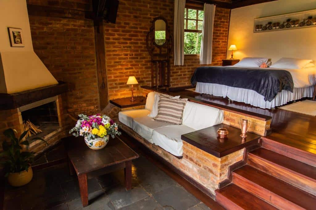 Quarto da Pousada das Araras. Uma cama de casal está em uma parte mais alta do ambiente, no lado direito. Abaixo dela, depois de uma escadinha, está um sofá, uma mesa de centro e uma lareira.