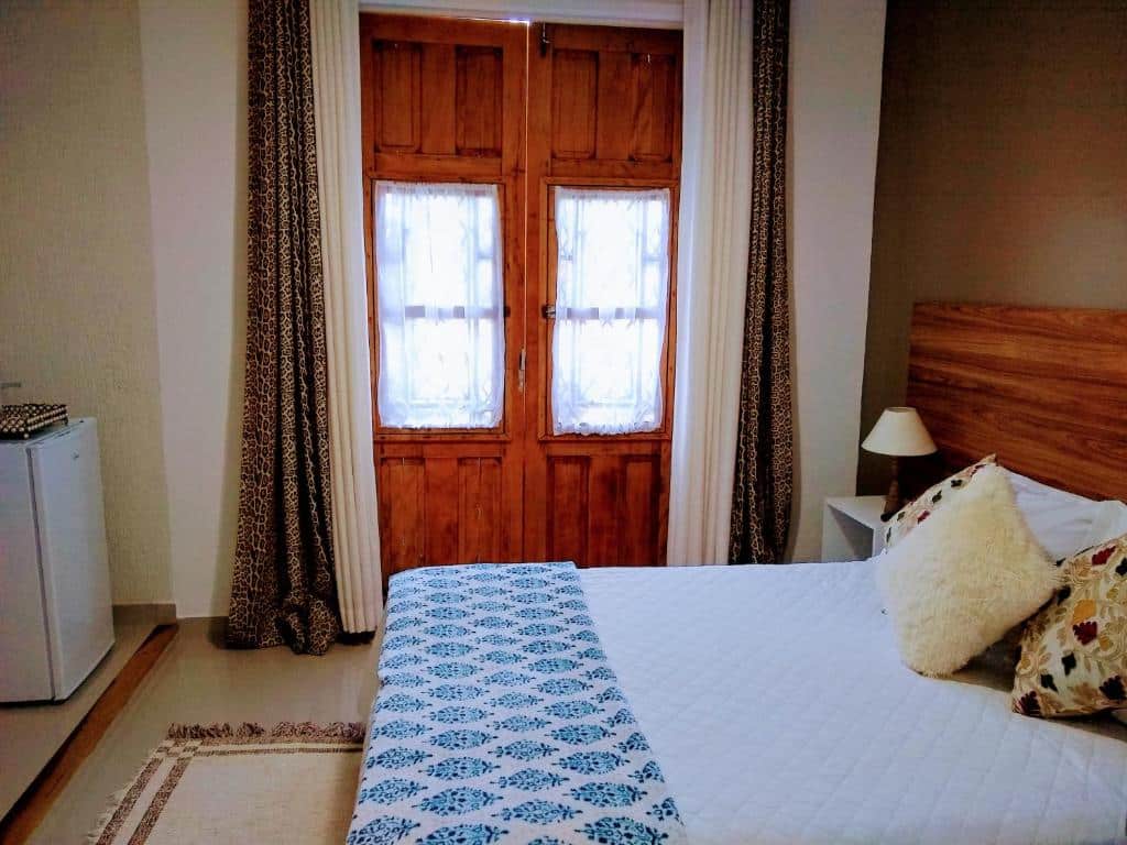 Quarto da Pousada do Lago com uma cama de casal com roupa de cama estampada, cabeceira de madeira, geladeira pequena e portas francesas com cortinas transparentes.