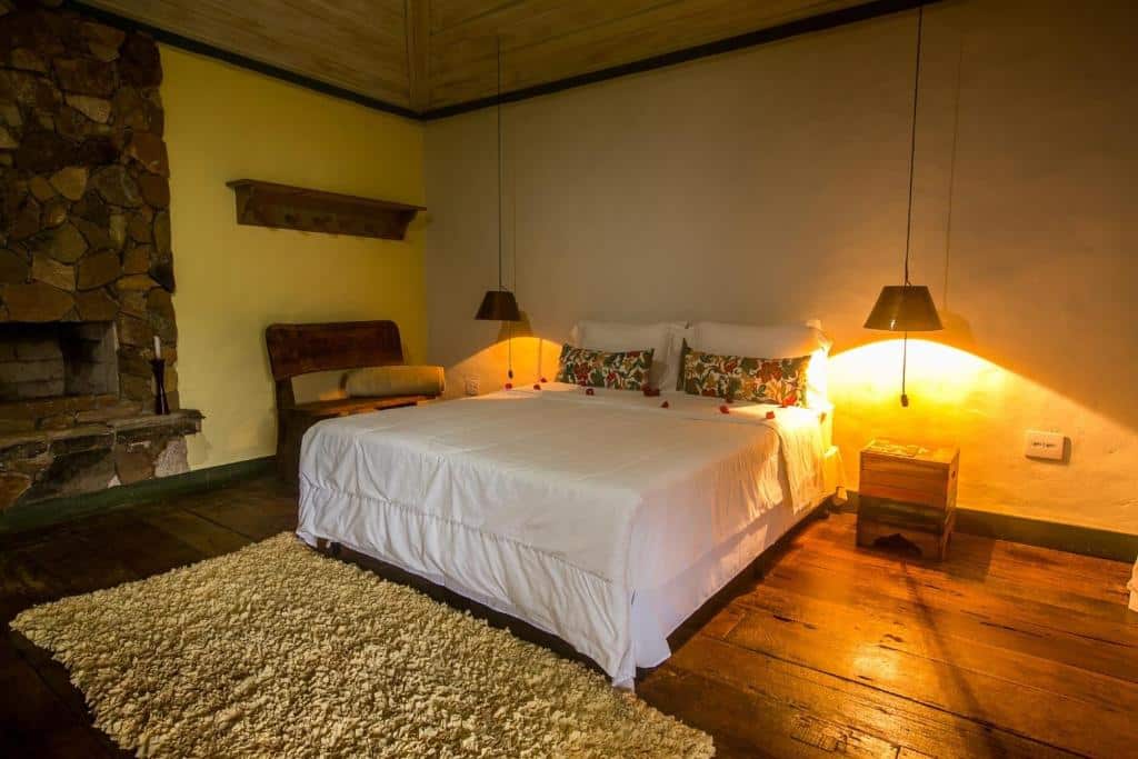 Quarto da Pousada Meu Recanto que mostra uma cama de casal ao centro e de cada lado da cama uma mesa de cabeceira e um lustre pendurado com luz amarela. Do lado esquerdo do quarto tem um banco de madeira e uma lareira de pedras. No quarto tem um tapete e o chão é de madeira. Imagem ilustrando post O que fazer em Ibitipoca.