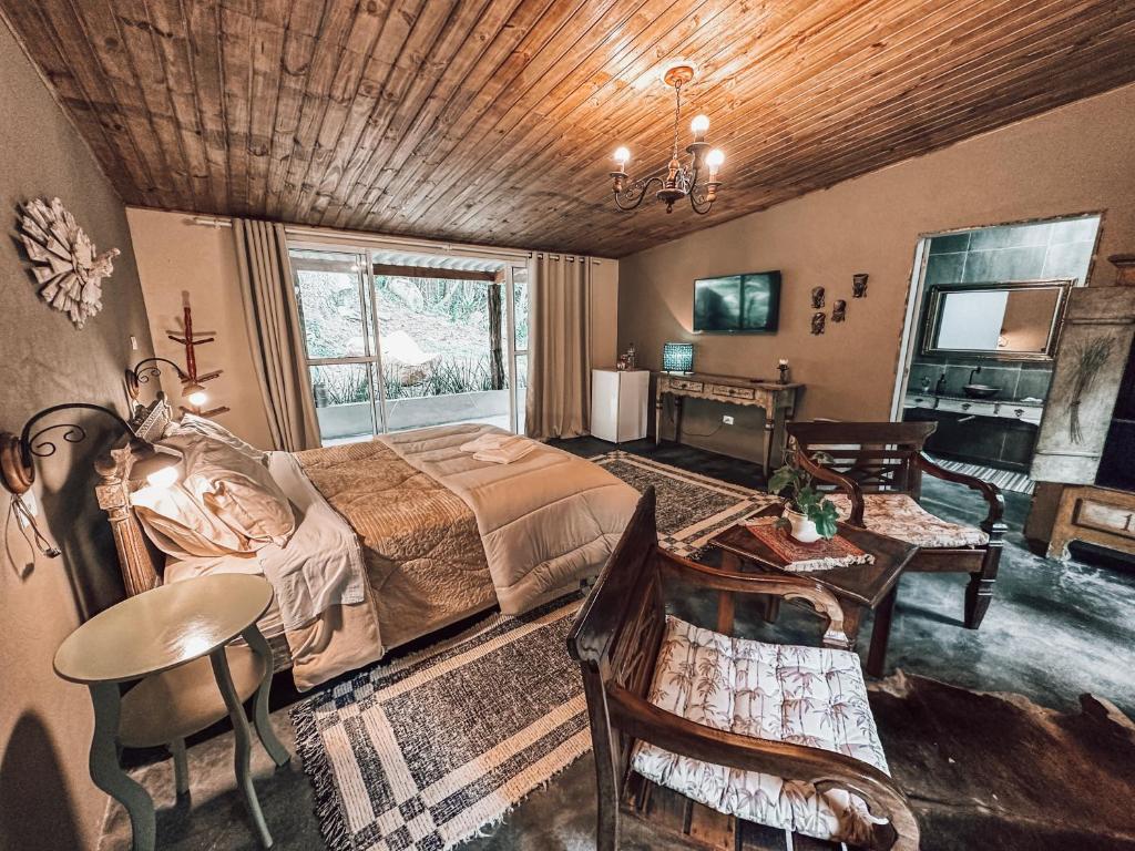 Quarto da Pousada Santuário Acalanto. Do lado direito duas poltronas de madeira com uma mesa de centro, no fundo a porte do banheiro. Do lado direito uma mesa de cada lado da cama de casal com um lustre, no fundo a varanda. De frente para a cama uma estante, um frigobar e uma televisão.