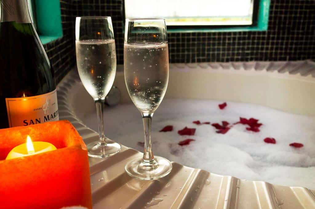 Banheira de hidromassagem da Pousada Vila Santa Barbara. Ela está cheia e coberta com algumas pétalas de rosa. No canto estão duas taças de champagne, a garrafa e uma vela.
