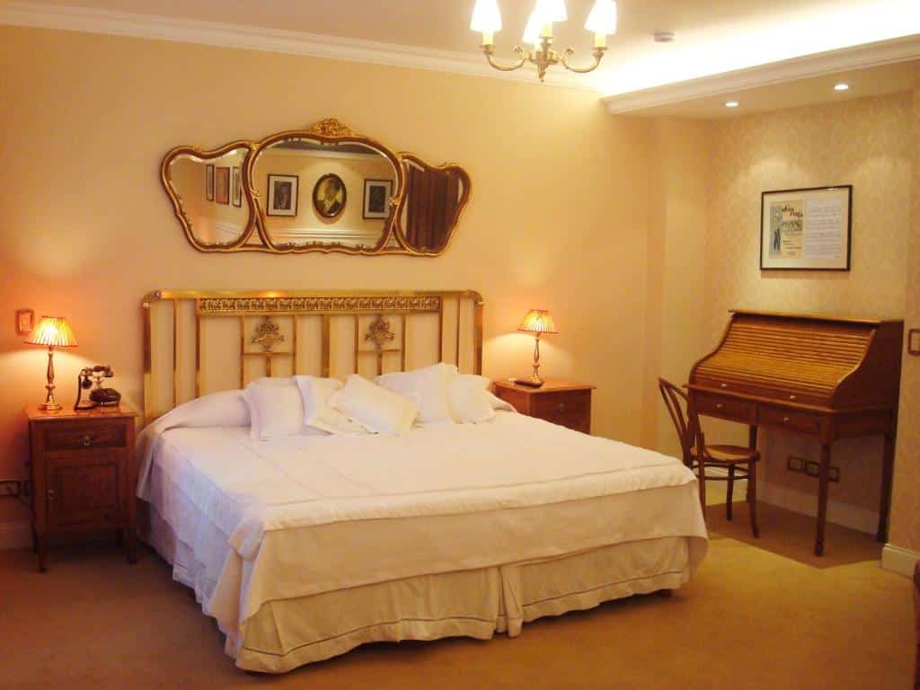 quarto do Tanguero Hotel Boutique Antique, um dos hotéis românticos em Buenos Aires, com cama de casal, mesinha e luminária de ambos os lados, mesa com cadeira em madeira, espelho em forma de maçã cortada, e detalhes clássicos