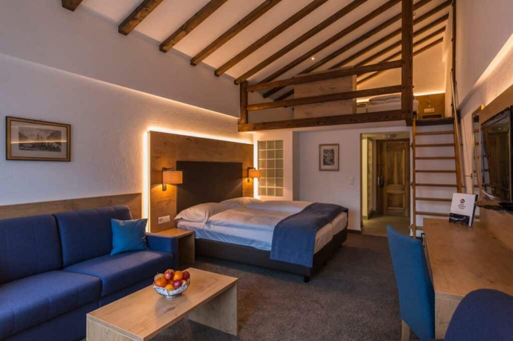 Quarto do Alpen Resort & Spa com uma sala de estar a frente com sofá e uma mesa quadrada do lado esquerdo e em frente a sala de estar uma mesa com cadeira e uma TV presa na parede, já ao fundo da área de estar uma cama de casal. Representa onde ficar em Zermatt.