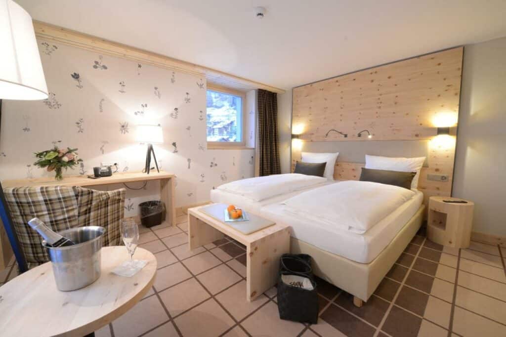 Quarto do Alpenhotel Fleurs de Zermatt com cama de casal do lado direito da imagem no centro do quarto com uma cômoda do lado esquerdo da cama. No pé da cama uma cômoda de madeira em frente a cama uma mesa redonda com bande de champanhe e uma taça e do lado direito da cama uma mesa de trabalho com cadeira. Representa onde ficar em Zermatt.