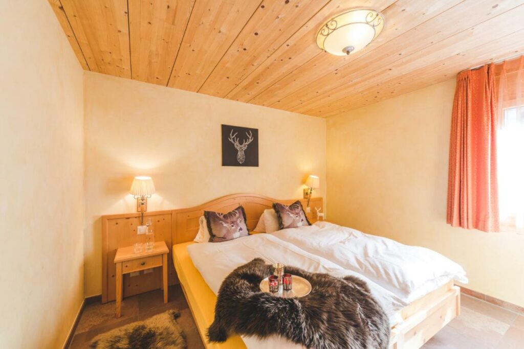 Quarto do Apartment Alpharmonie com cama de casal do lado esquerdo da imagem no centro do quarto com dias cômodas em cada lado da cama com luminária em cima da cama uma bandeja com três copos. Representa onde ficar em Zermatt.