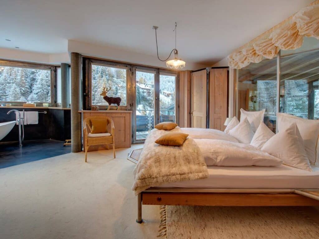 Quarto do Backstage Hotel Serviced Apartments com cama de casal do lado direito da imagem do lado direito da cama uma poltrona e cômoda, além de uma porta de vidro que dá acesso a varanda. Representa onde ficar em Zermatt.