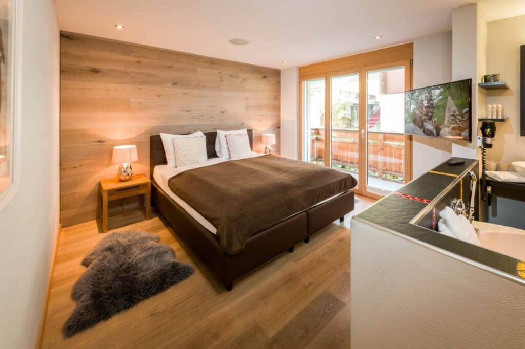 Quarto da Casa Della Luce Apartments com cama de casal do lado esquerdo da imagem no centro do quarto com uma cômoda em cada lado da cama com luminária e do lado esquerdo da cama acesso a varanda por uma porta de vidro e no canto uma TV presa. Representa onde ficar em Zermatt.