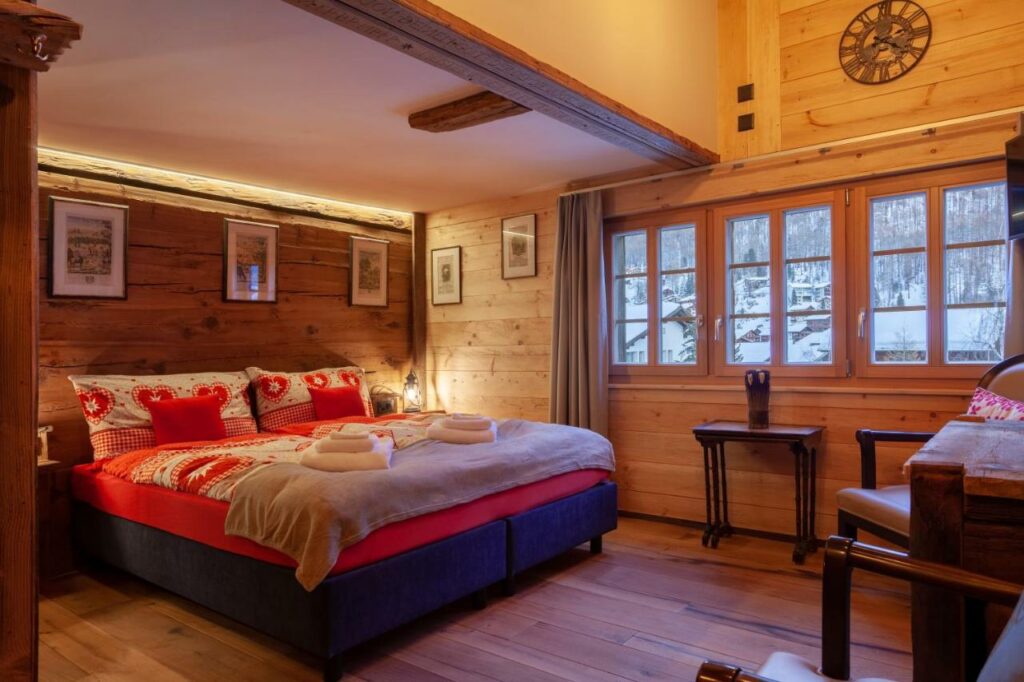 Quarto do Chalet Schuler com uma cama de casal do lado esquerdo da imagem em frente a cama duas cadeiras e uma mesa. Representa onde ficar em Zermatt.