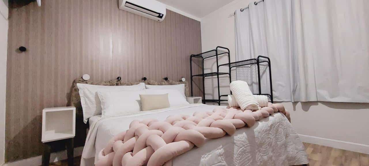 A imagem mostra o quarto do Apto Essepê, um dos airbnb em São Paulo, com paredes texturizadas em tons de marrom claro e um piso de madeira. Há uma cama de casal com roupa de cama branca, uma travesseira longa em forma de trança rosa claro sobre a colcha, e almofadas decorativas. Ao lado da cama, há um criado-mudo branco e, acima, um ar-condicionado. Contra a parede oposta à cama, há uma estante metálica preta e ao lado uma cortina branca esticada na parede.