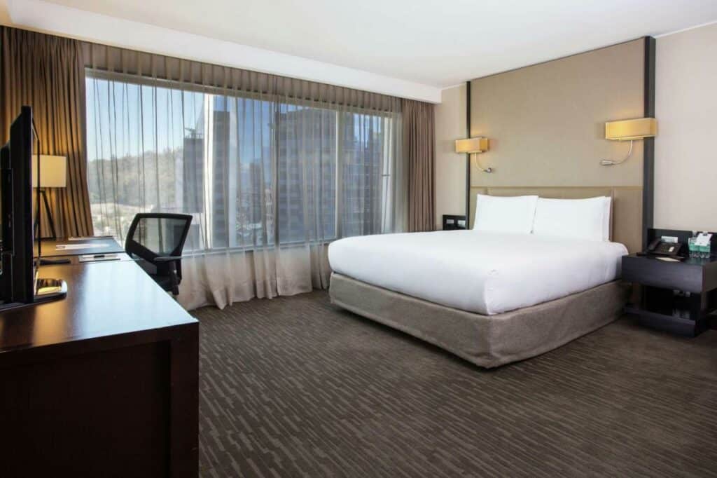 Quarto do DoubleTree by Hilton Santiago – Vitacura com cama de casal do lado direito da imagem no centro e uma cômoda em cada lado da cama e em frente a cama uma cômoda com TV em cima. Representa hotéis no Bairro Las Condes em Santiago.