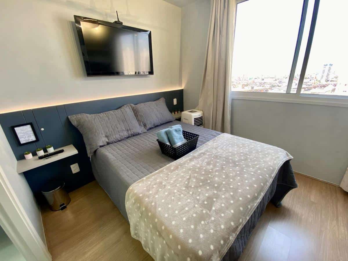 Quarto do Fantástico Apartamento Em Frente Ao Metrô Brás, sendo ele bem iluminado com uma grande janela que oferece vista para uma paisagem urbana. A cama de casal está no centro do quarto, com uma colcha cinza e uma manta branca com pequenos pontos cinza sobre ela. Há um painel azul escuro que serve de cabeceira, se estendendo ao longo da parede atrás da cama, com uma prateleira branca logo abaixo de uma televisão de tela plana que está montada na parede. Sobre a prateleira, há três velas verdes e controle de TV.
