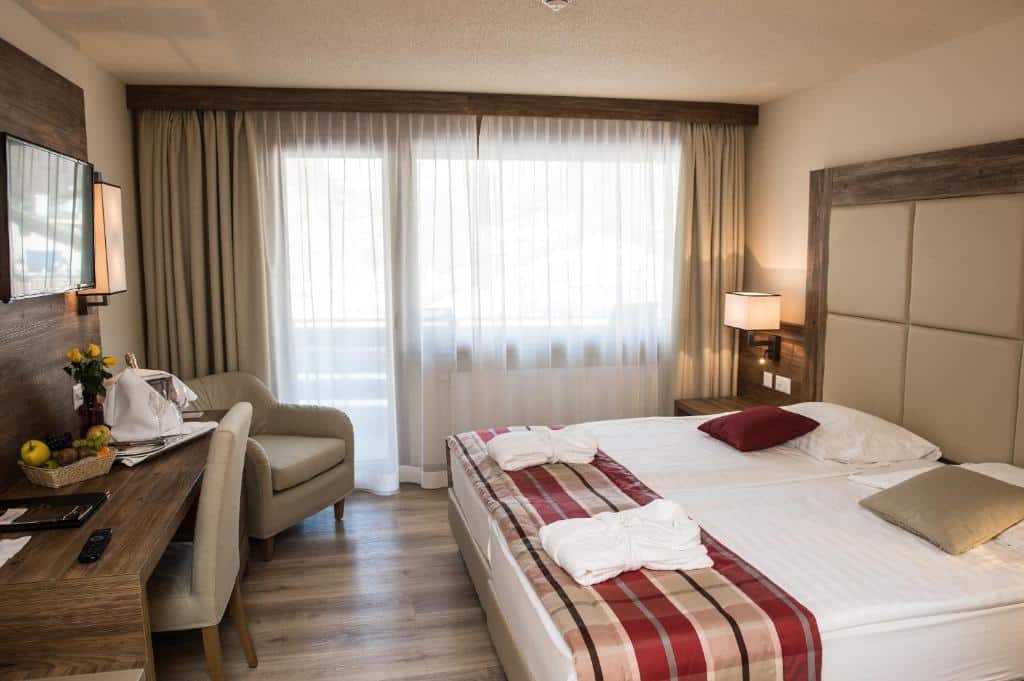 Quarto do Hotel Ambassador Zermatt com cama de casal do lado direito da imagem do lado direito da cama uma luminária presa na parede em frente a cama uma mesa com cadeira, uma TV presa na parede e uma poltrona. Representa onde ficar em Zermatt.