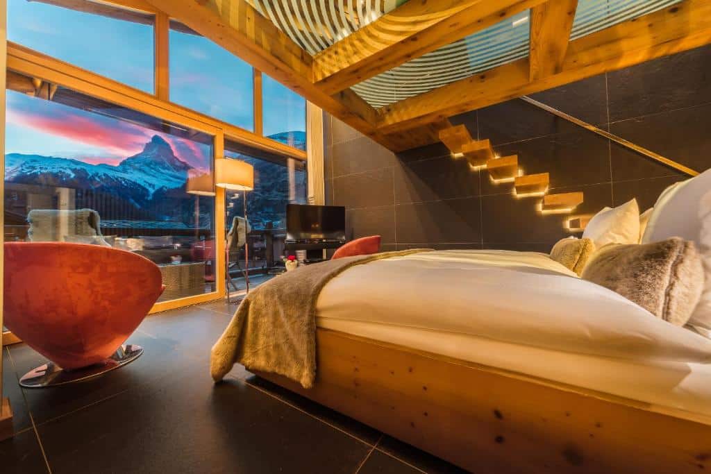 Quarto do Hotel Bellerive com uma cama de casal do lado direito da imagem do lado esquerdo da cama um poltrona e uma janela ampla com vista para as montanhas. Representa onde ficar em Zermatt.