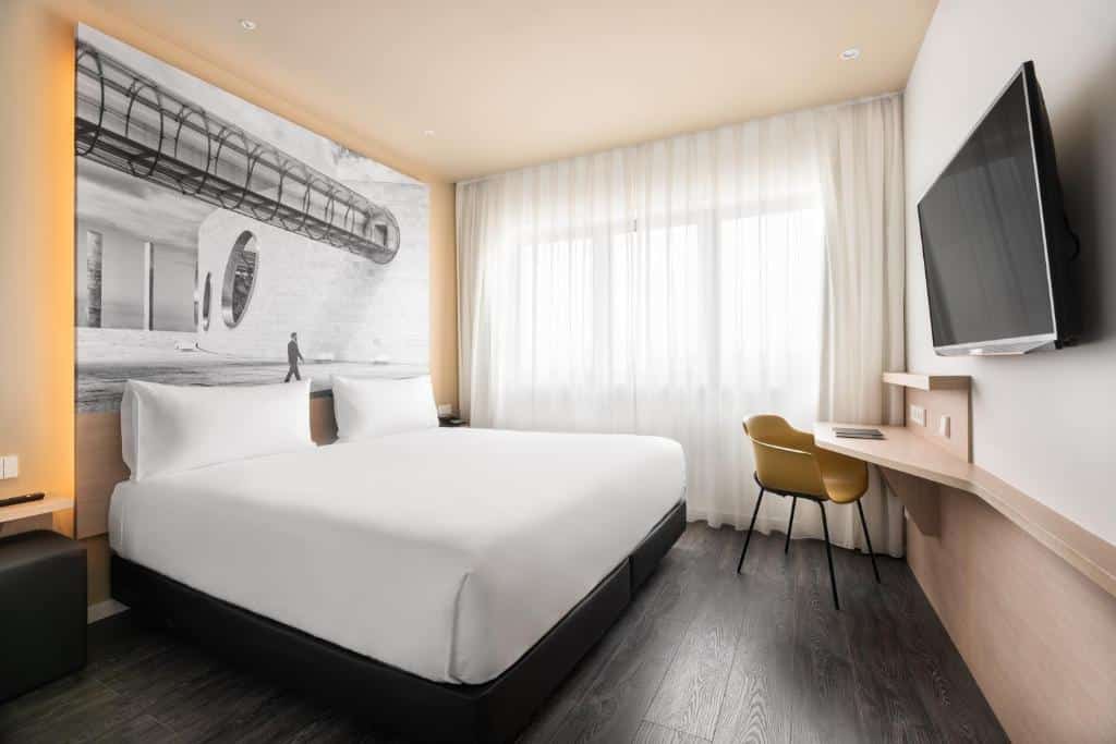 Quarto do Ikonik Lisboa com uma cama de casal, uma janela ampla com cortinas brancas, uma televisão de frente para a cama e uma pequena mesa de escirtório próxima da janela, junto de uma cadeira