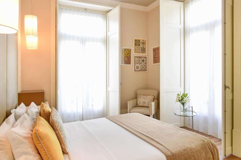 Quarto do My Story Hotel Ouro em tons de bege e branco, há duas sacadas iluminando o local, uma cama de casal com diversas almofadas, alguns quadros e luminárias nas paredes e uma poltrona estofada