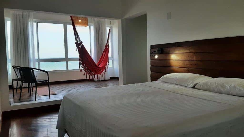 Quarto da Pousada Recanto Ha Mar com uma cama de casal e uma ampla janela com vista pro mar com uma rede de descanso pendurada próxima dela