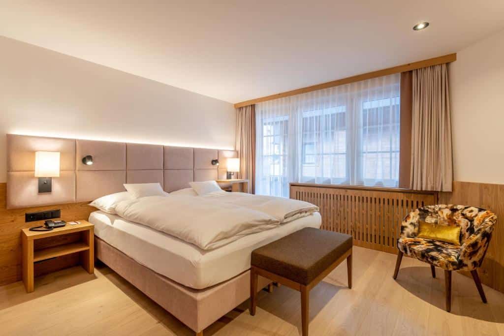 Quarto do Resort La Ginabelle com cama de casal do lado esquerdo da imagem no centro do quarto com uma cômoda em cada lado da cama no pé da cama um banco e do lado esquerdo uma poltrona. Representa onde ficar em Zermatt.