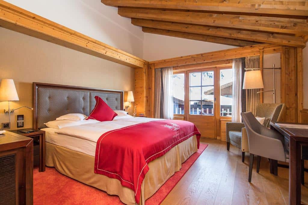 Quarto do Riffelalp Resort 2222m com cama de casal do lado esquerdo da imagem com uma cômoda em cada lado com luminárias e em frente a cama uma mesa com cadeira e do lado esquerdo da mesa uma poltrona. Representa onde ficar em Zermatt.