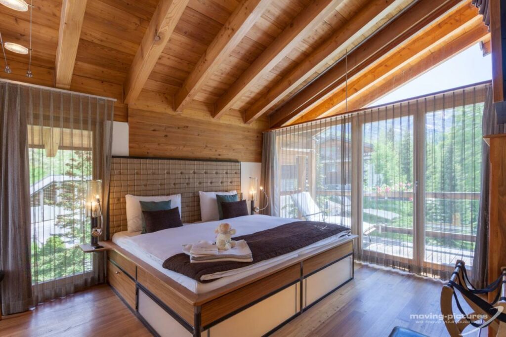 Quarto do Suitenhotel Zurbriggen com cama de casal do lado esquerdo da imagem no centro do quarto do lado esquerdo da cama uma porta de vidro coberta com cortinas que dá acesso a varanda. Representa onde ficar em Zermatt.
