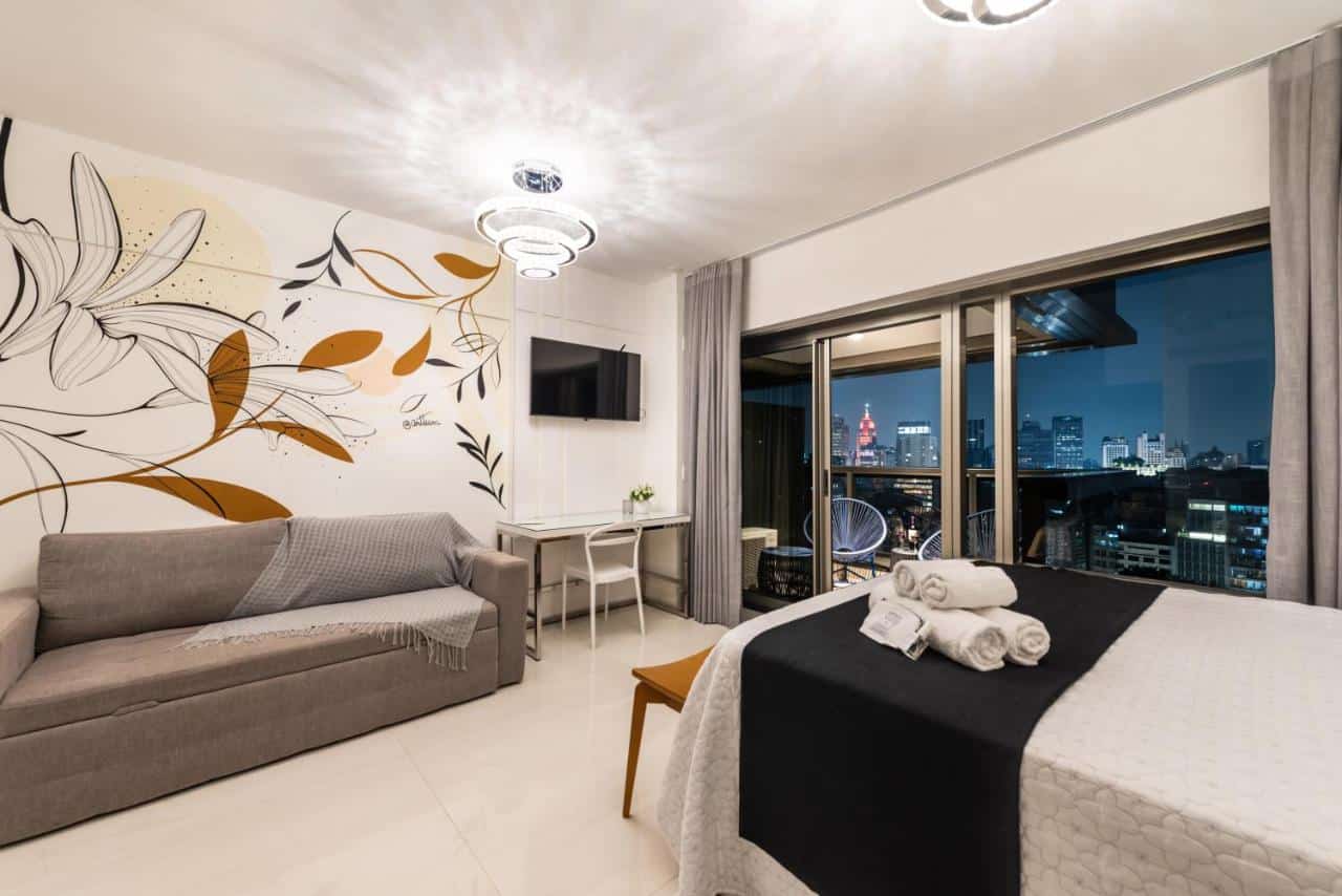 Quarto de um dos apartamentos da Romemari's Apartments República, um dos airbnb em São Paulo. A foto mostra um estúdio com grandes janelas de vidro oferecendo uma vista noturna da cidade. Há uma cama de casal à direita com uma colcha branca e coberta preta no pé da cama, acompanhada de toalhas dobradas e itens de higiene pessoal. À frente da cama, uma mesa de trabalho branca com uma cadeira e um vaso de plantas. À esquerda, há um sofá cinza com uma manta e ao fundo uma grande pintura mural de flores estilizadas em tons de branco, preto e dourado. Acima, uma luminária de teto emite uma luz suave, e uma TV de tela plana está montada na parede, à frente da cama. O piso é de azulejo branco, e o espaço é moderno e aconchegante.