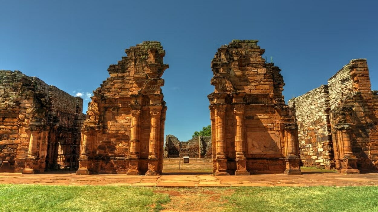 A imagem mostra uma fachada de pedras esculpidas, com detalhes arquitetônicos impressionantes das Ruínas de San Ignacio Miní. O céu azul e o gramado verde ao redor realçam a beleza histórica do local.