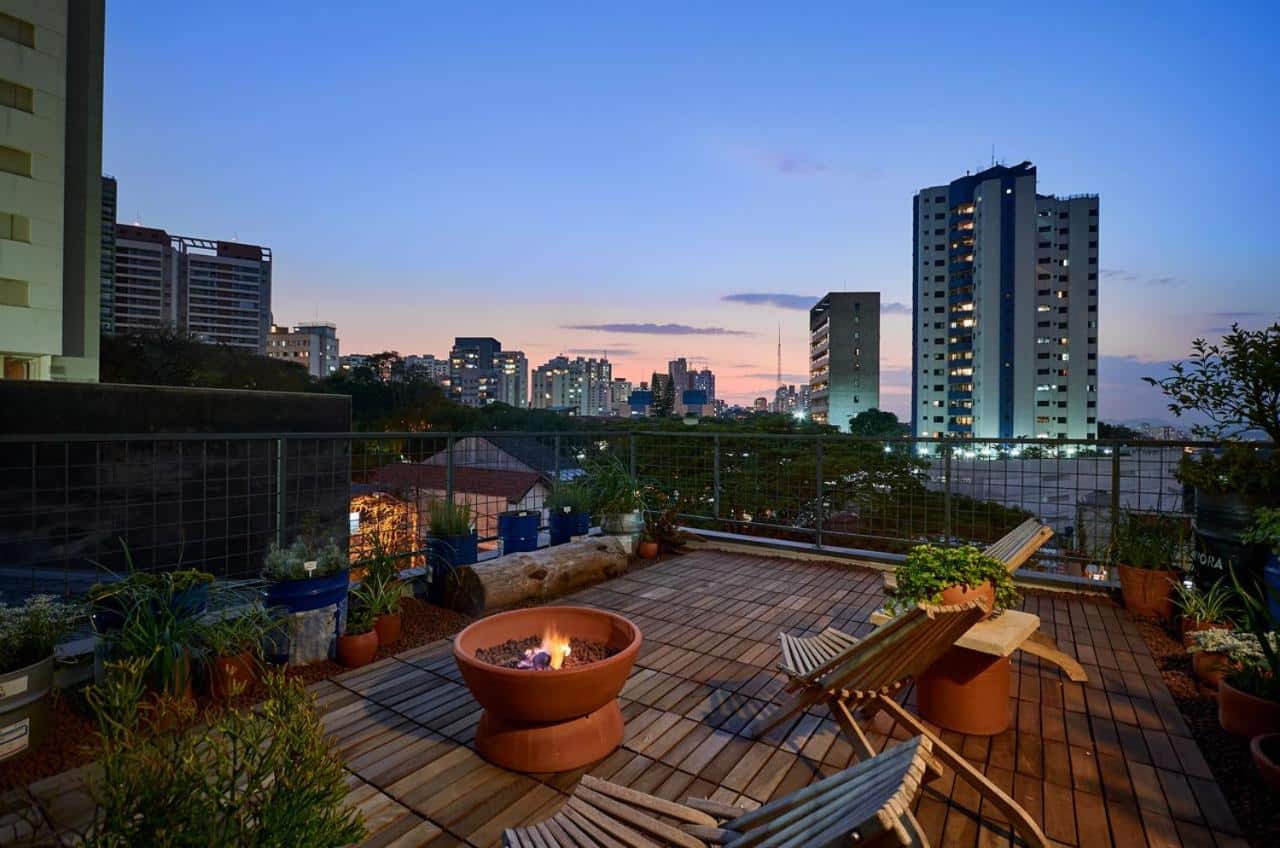 A foto captura o terraço urbano ao entardecer da Vila Pitanga Hosting, um dos airbnb em São Paulo. No centro, há uma fogueira de chão em cerâmica com fogo aceso, emitindo um brilho acolhedor. À volta da fogueira, dispostos sobre um deck de madeira, estão dois bancos de madeira e várias plantas em vasos de diferentes tamanhos e estilos. Ao fundo, o skyline da cidade com prédios de diversos tamanhos se destaca sob um céu colorido pelo pôr do sol. O ambiente transmite uma sensação de tranquilidade e refúgio no meio urbano.