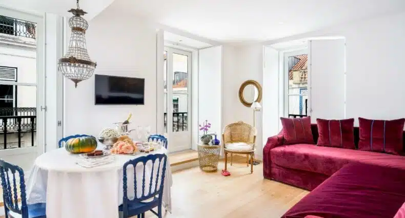 Sala de estar ampla do Residentas Aurea com sofás, uma pequena sacada, uma televisão e uma mesa com café da manhã e quatro lugares, além de itens de decoração dourados, para representar airbnb em Lisboa