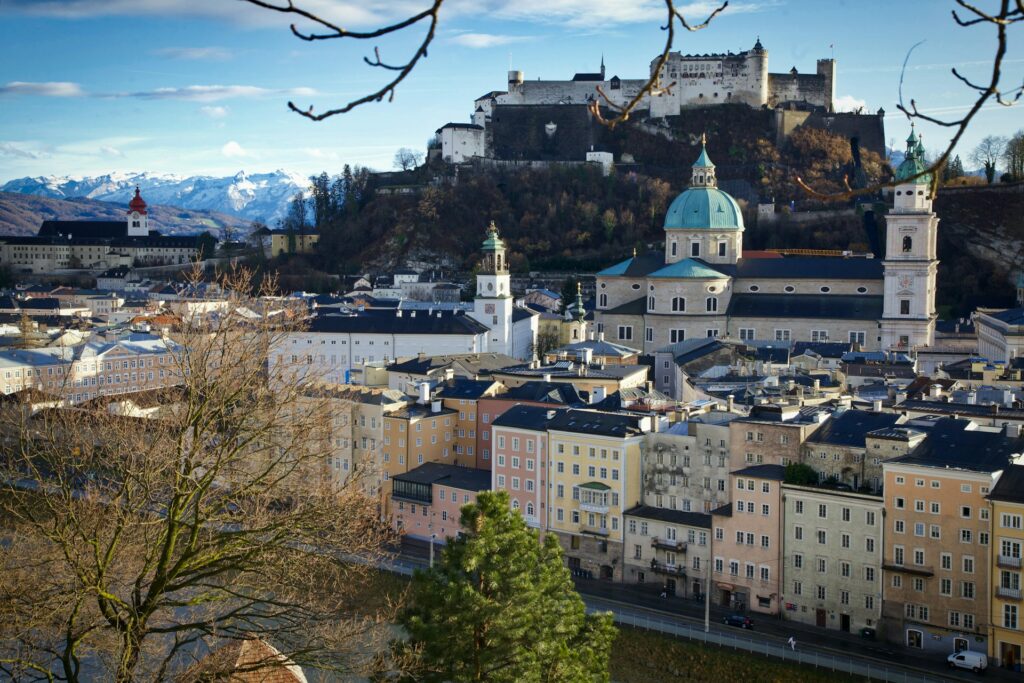 Vista panorâmica da cidade de Salzburg, Áustria, emoldurada por um céu azul claro e montanhas cobertas de neve ao fundo. Destaca-se na imagem o imponente Forte de Hohensalzburg, uma fortaleza medieval situada no topo de uma colina. Abaixo, a cidade é um mosaico de edifícios coloridos e igrejas barrocas com cúpulas e torres proeminentes. A composição é realçada pela luz natural que ilumina a cidade.