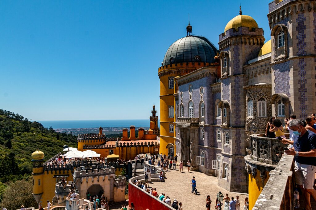 Vista do Palácio da Pena em Sintra, Portugal. O palácio destaca-se por sua arquitetura romântica com cúpulas adornadas em tons de amarelo e azulejos intricados. Turistas passeiam e apreciam a vista no terraço espaçoso, que oferece uma vista panorâmica sobre a vegetação dos arredores e o mar ao fundo. O céu claro e o dia ensolarado acentuam as cores brilhantes e detalhes arquitetônicos deste marco português.