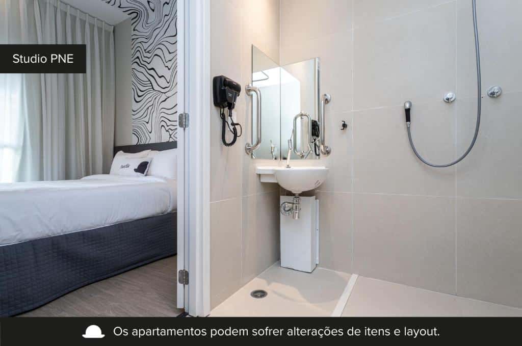 A imagem mostra o interior do Estúdio PNE do House of Charlie, um dos airbnb em São Paulo. Ele é adaptado para acessibilidade. À esquerda, há uma cama com cabeceira e roupa de cama em tons de branco e azul escuro, e acima, uma cortina cinza. À direita, vemos o banheiro com piso e paredes de azulejos bege. Um espelho está acima de um lavatório branco com torneiras acessíveis e barras de apoio, e ao lado, uma ducha. Há uma mensagem na parte inferior da imagem indicando que os itens e o layout dos apartamentos podem sofrer alterações.