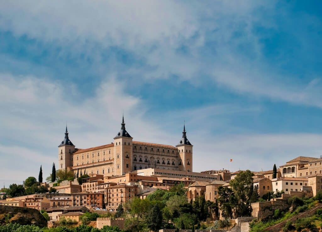 O imponente Alcázar de Toledo, de cor creme e com torres de telhado azulado, domina a paisagem, situado no topo de uma colina com vistas para a cidade antiga abaixo, repleta de edifícios históricos. O céu azul com nuvens esparsas proporciona um fundo dramático, enquanto a vegetação verde adiciona um contraste natural à arquitetura de pedra.