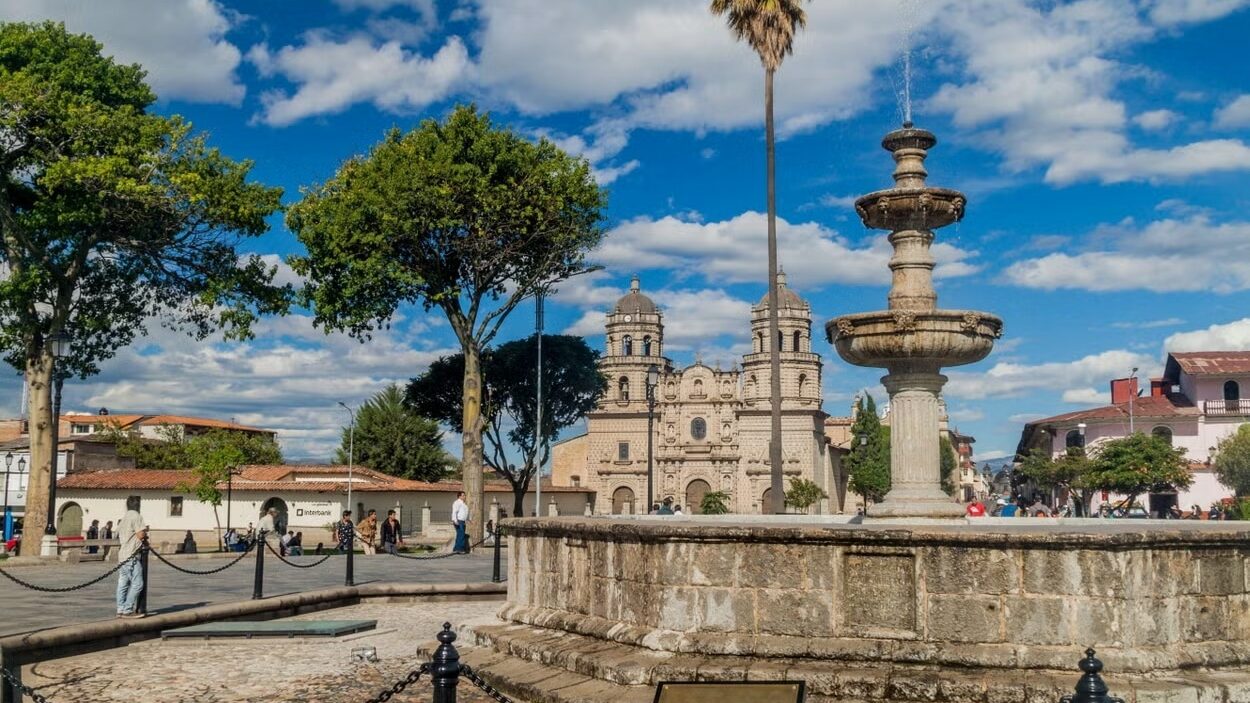 Praça das Armas de Cajamarca, com um chafariz cercado por uma corrente preta, árvores e um prédio histórico ao fundo. A imagem foi tirada durante um dia ensolarado, com poucas núvens no céu azulado. Foto: Civitatis